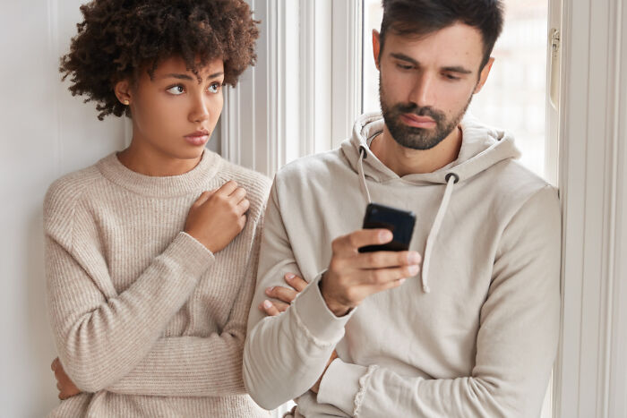 A woman looks suspiciously at a man engrossed in his phone, representing disturbing things people learn about friends and seeing them differently.