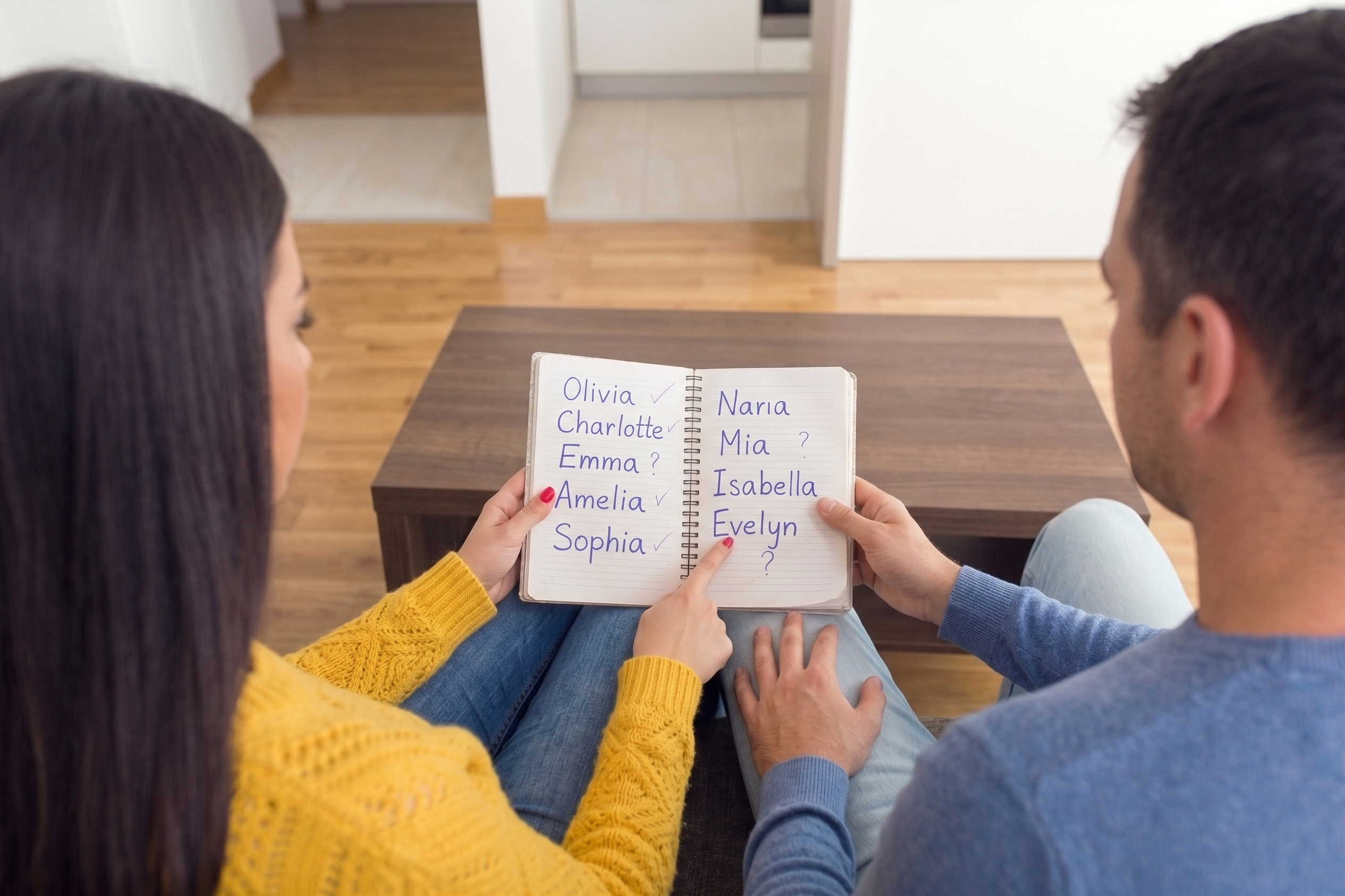 A couple sits on a couch, looking at a notebook with baby names. The husband wants to honor his mom, the wife wants to honor her.