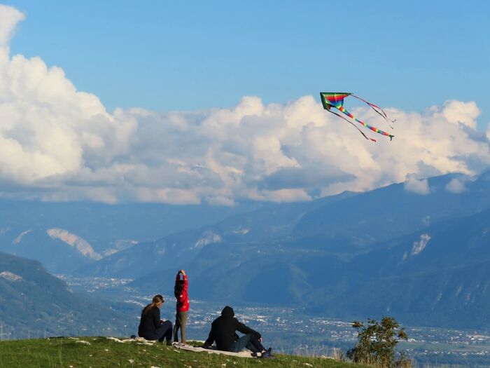 Three strangers fly a colorful kite on a hill overlooking a valley, showcasing the "Like A Painting" photo trend.