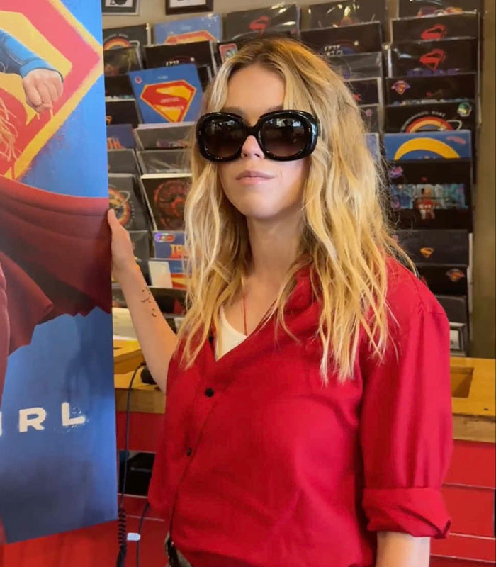 Sydney Sweeney posing in sunglasses next to a Supergirl movie display with records in the background.