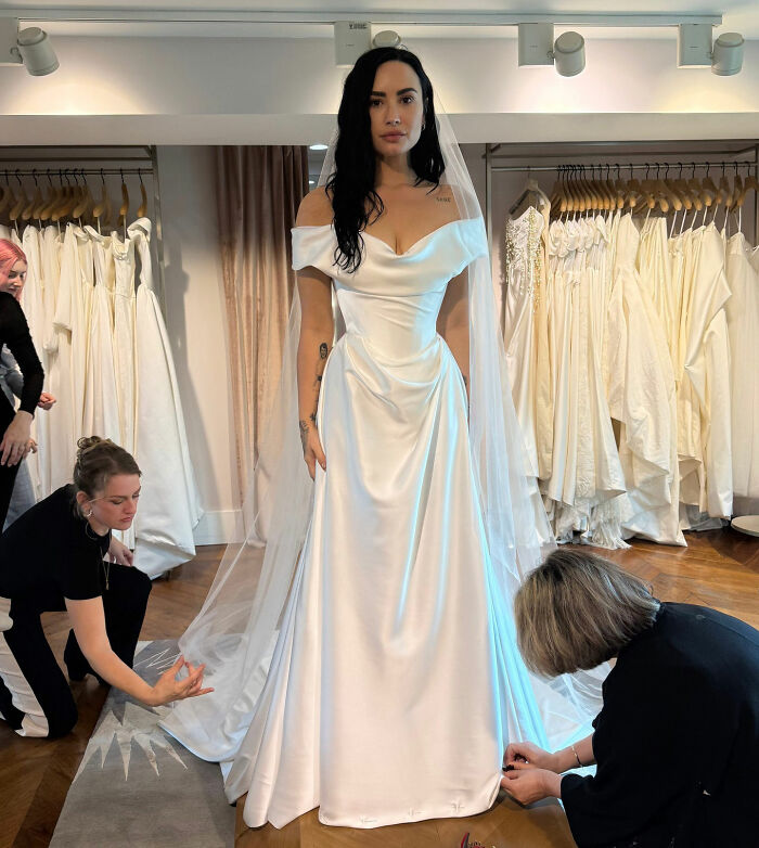 A celebrity in a stunning wedding dress, with a long veil being adjusted by two assistants.