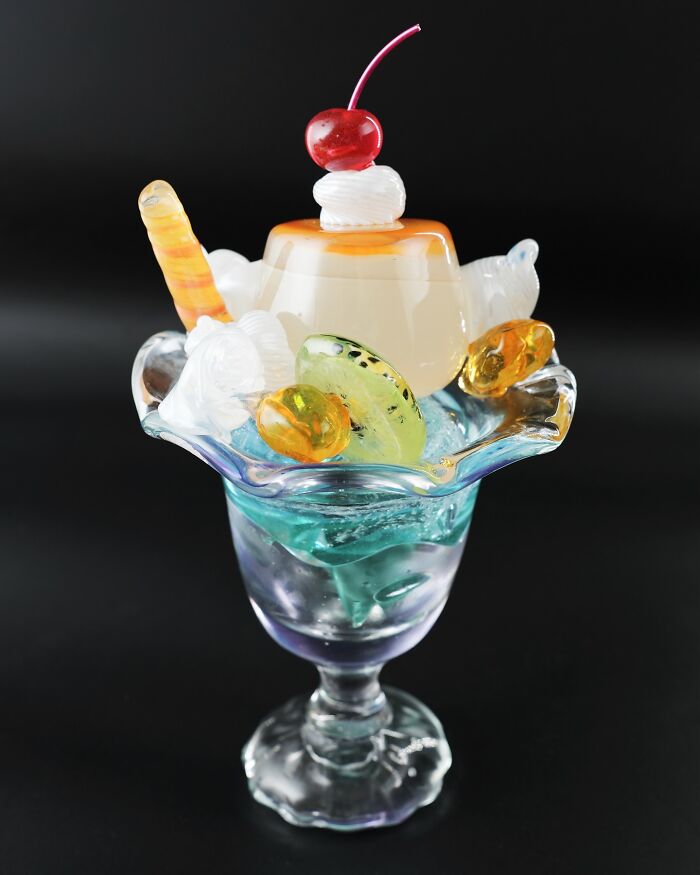 A hyper-realistic glass food parfait with pudding, fruit slices, and a cherry on top in a glass dish.
