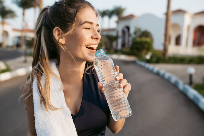 Happy woman drinking water outside during workout with towel on shoulder