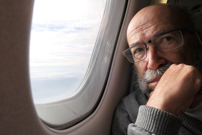 Elderly man with glasses looking out of a plane window during a flight, showing a plane interaction.