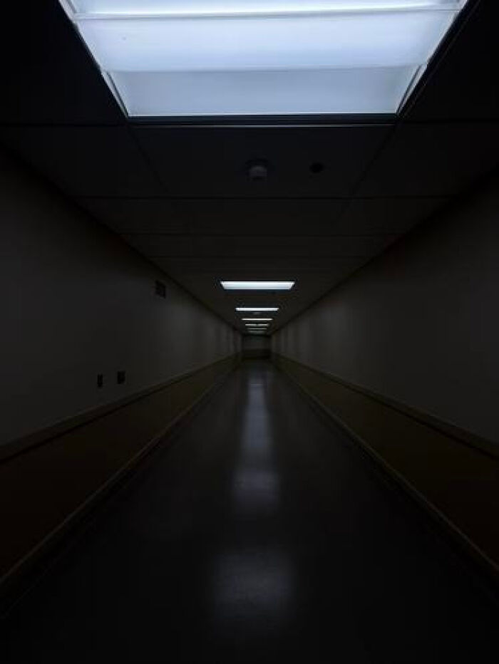 A long, dark hospital-like corridor with dim fluorescent lights, creating an unsettling liminal space.
