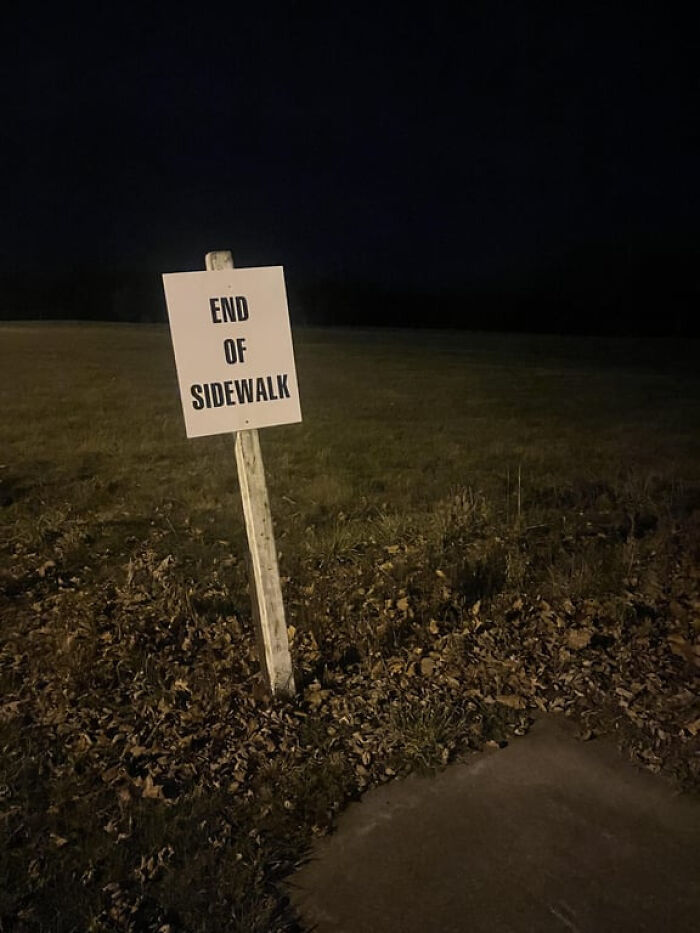 An unsettling liminal space: a white 'END OF SIDEWALK' sign in a dark, grassy field at night.