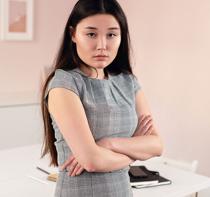 A woman with long dark hair, wearing a grey dress, looks sternly at the camera with her arms crossed, discussing her messy hair and job.