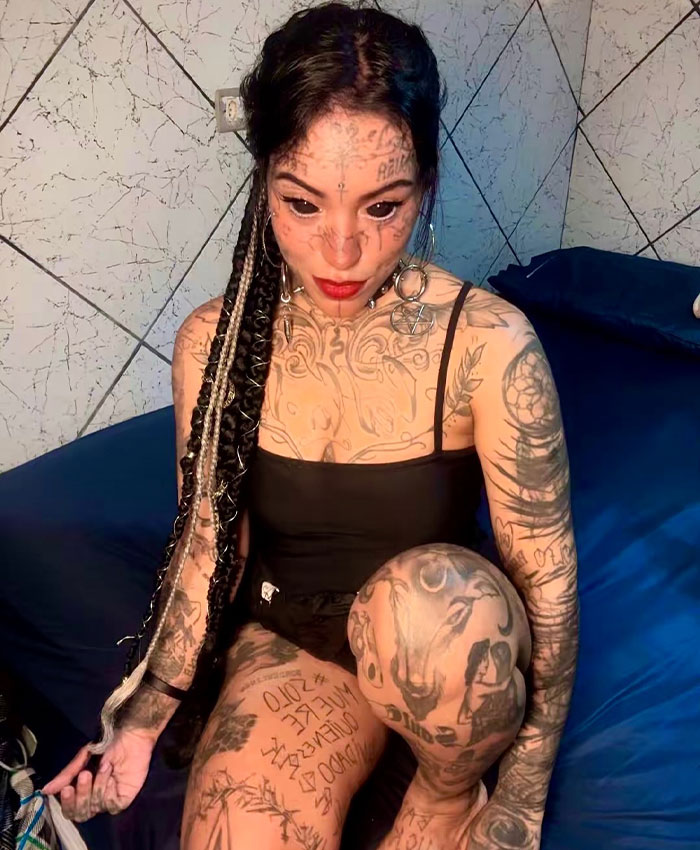 Woman with tattooed eyes sitting on a bed, showing her full body tattoos.