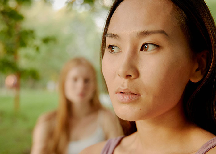 A woman looking away with another woman blurred in the background, showing things close friends commit that broke people's trust forever.