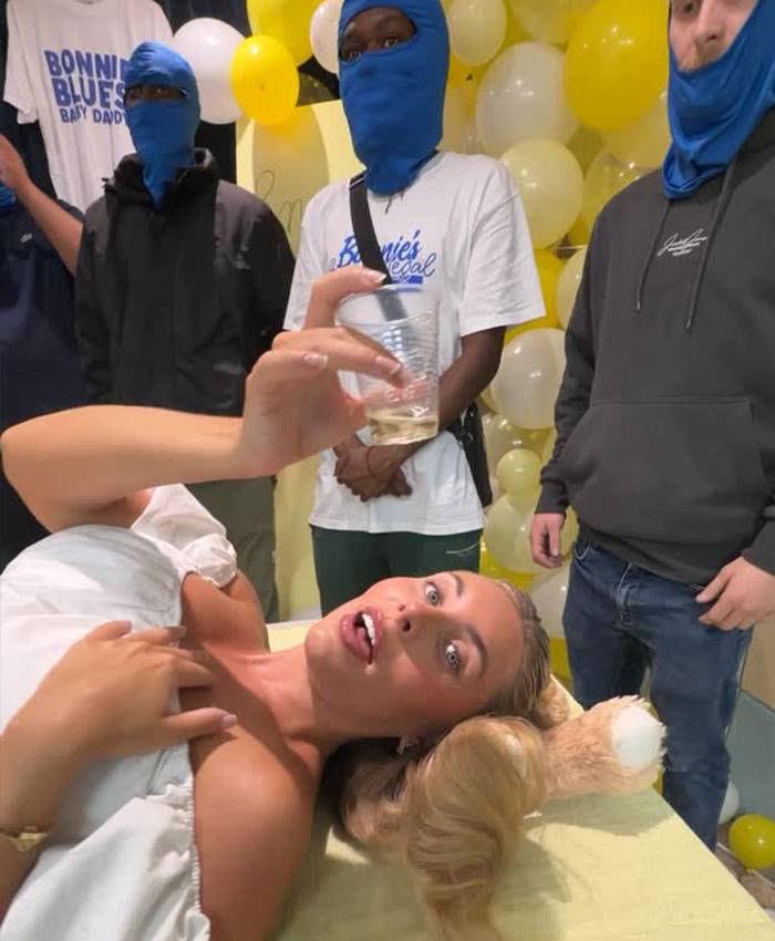 Bonnie Blue reclines, holding a drink, with friends in blue masks around her, enjoying her baby shower celebration.