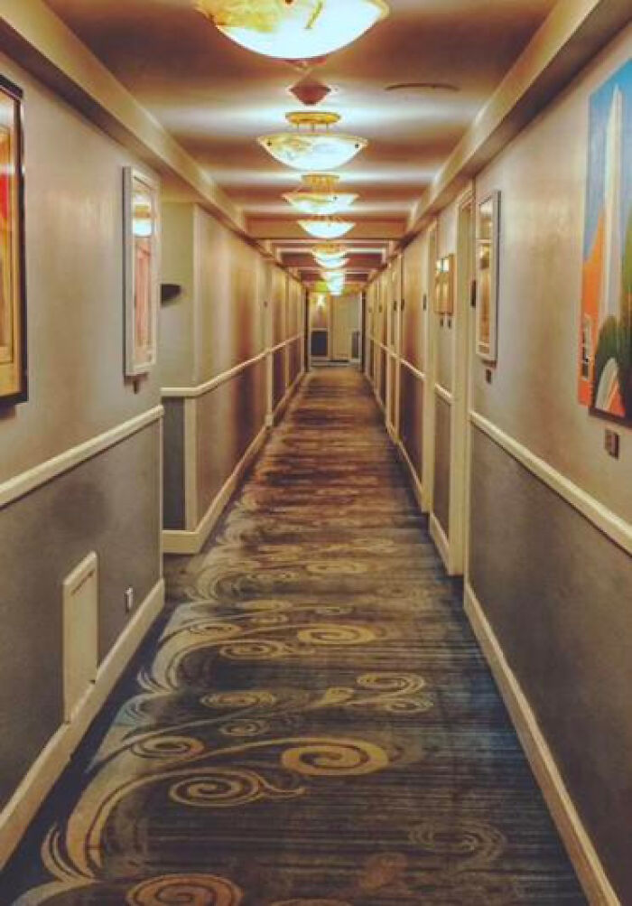 A long, empty hotel hallway with patterned carpet and overhead lights, a haunting liminal space.