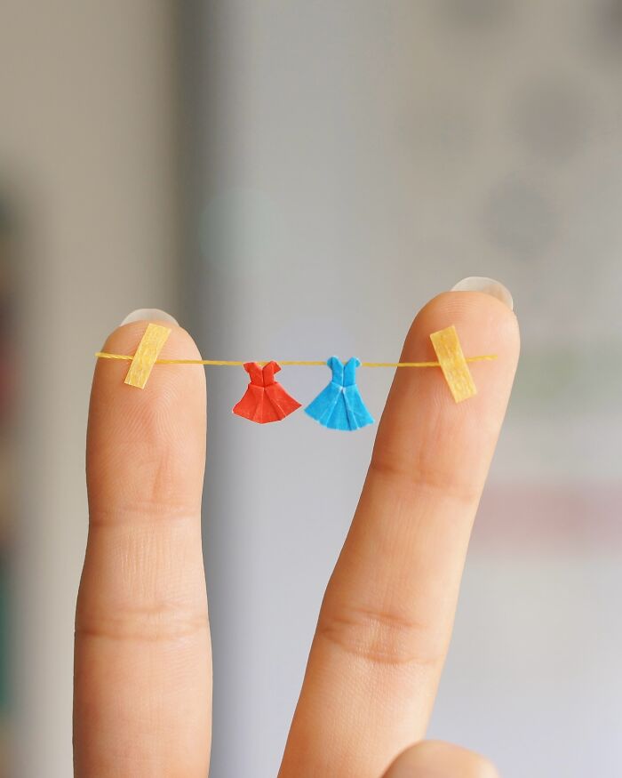 Two fingers holding a string from which two miniature paper figures of dresses, one red and one blue, are hanging like laundry.
