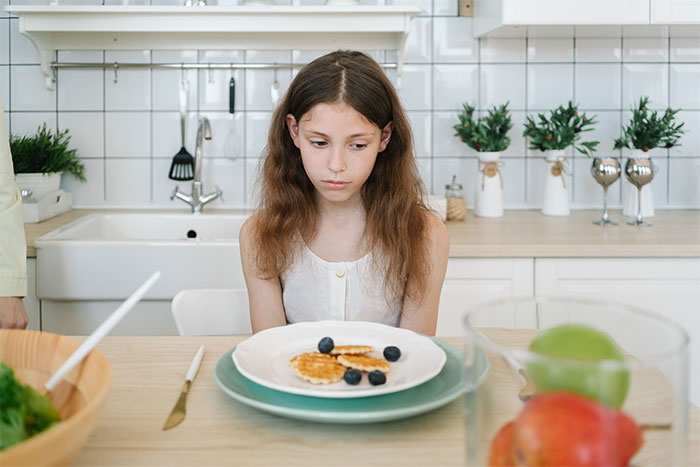A young girl looking sad at a plate of food, highlighting a kid's food issue.
