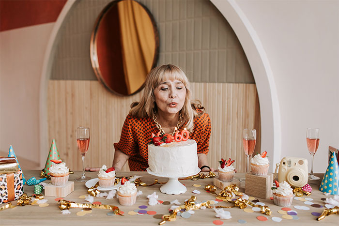 A woman blowing out 60th birthday cake candles, surrounded by cupcakes and champagne glasses, suspecting why someone got sick.