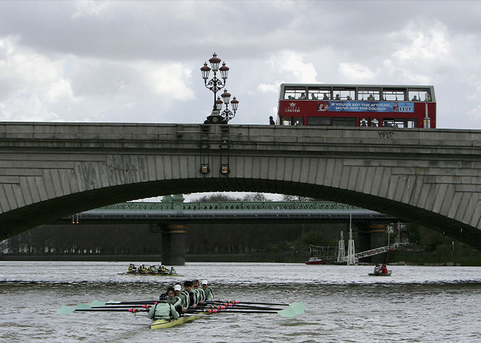 A red double-decker bus on a bridge over a river with crew boats below, related to the jogger pushing woman incident. A red double-decker bus on a bridge over a river with crew boats below, related to the jogger pushing woman incident.