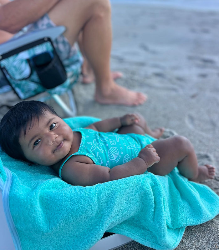A baby, born after an IVF mix-up, lies on a blue towel on the beach, smiling up at the camera. A baby, born after an IVF mix-up, lies on a blue towel on the beach, smiling up at the camera.