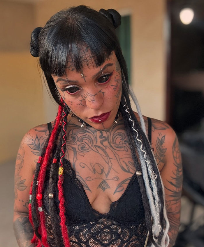 Woman with tattooed eyes and dreadlocks in black top, revealing her body and facial tattoos.