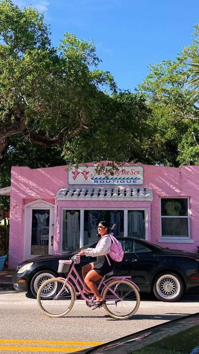 A woman on a pink bicycle in front of a pink boutique, part of the wholesome "Like A Painting" photo trend.
