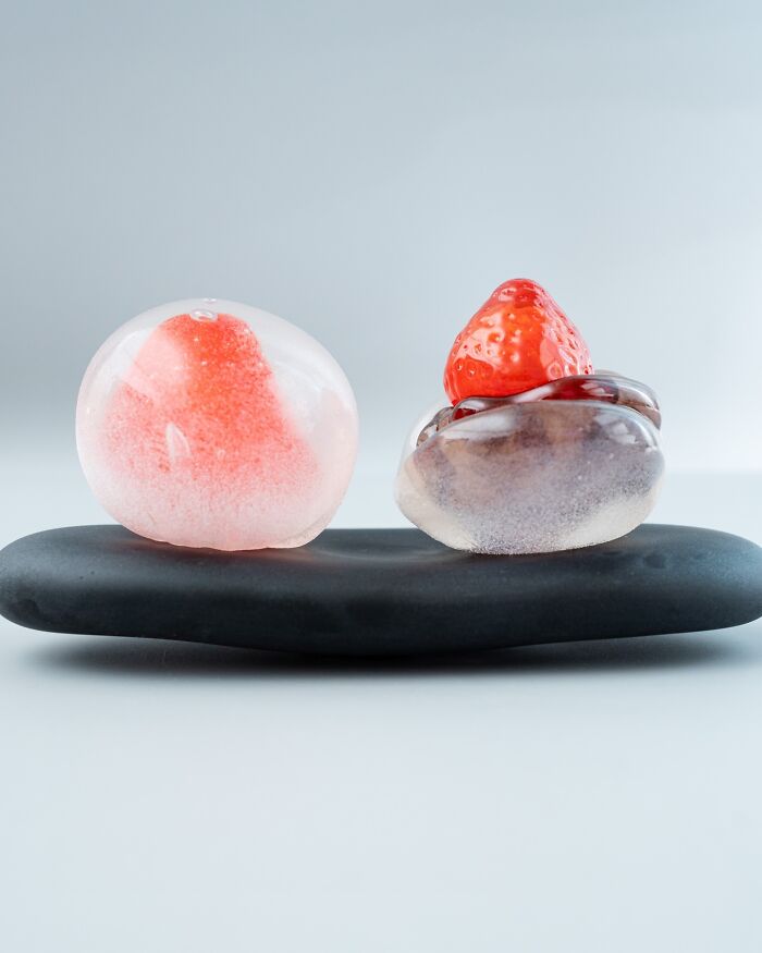 Two pieces of hyper-realistic glass food resembling desserts with strawberries, on a black platter.