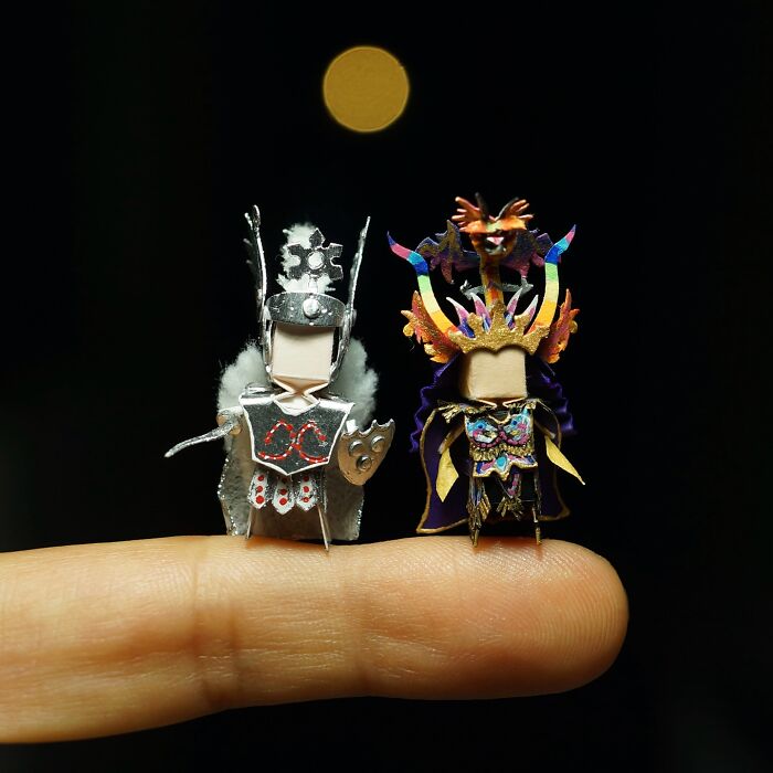 Two intricately detailed miniature paper figures of warriors standing on a fingertip.
