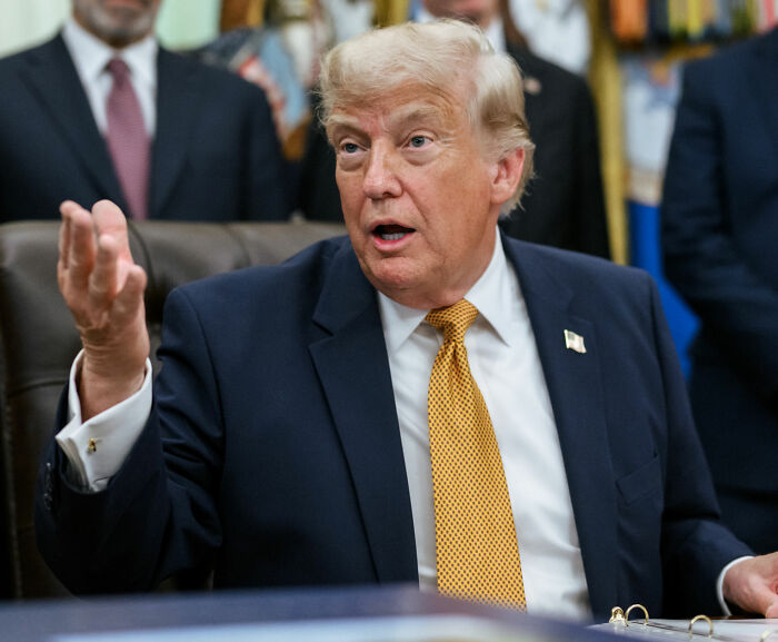Donald Trump gesturing while speaking, seated at a desk.