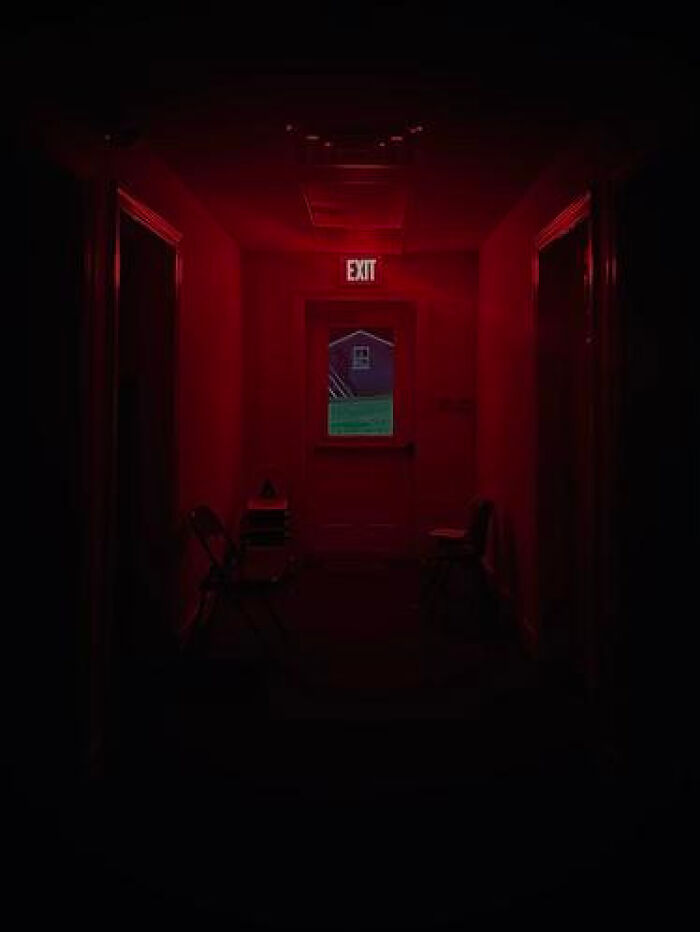 A dark, red-lit hallway with an illuminated EXIT sign above a door leading to a green outdoor liminal space.