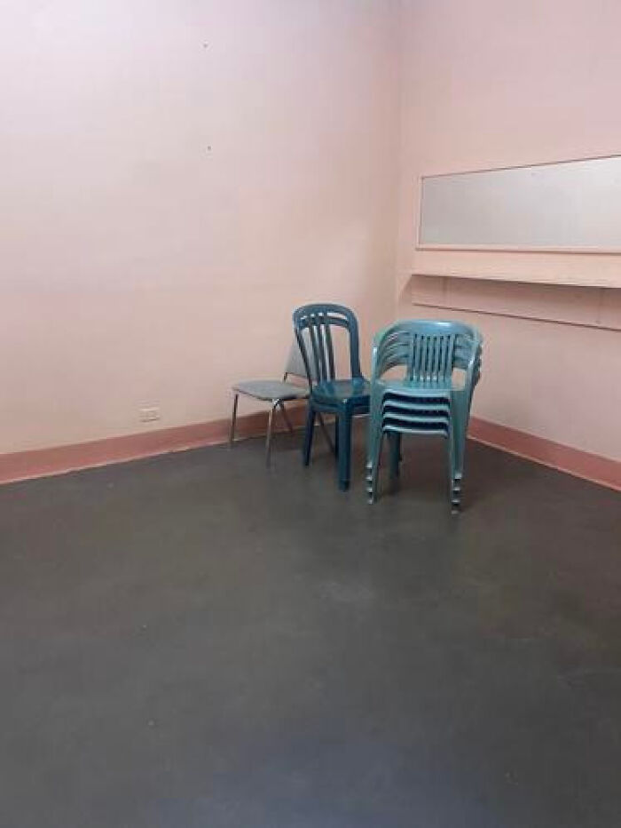 A haunting liminal space with a single chair and a stack of chairs against a pale pink wall and dark floor.