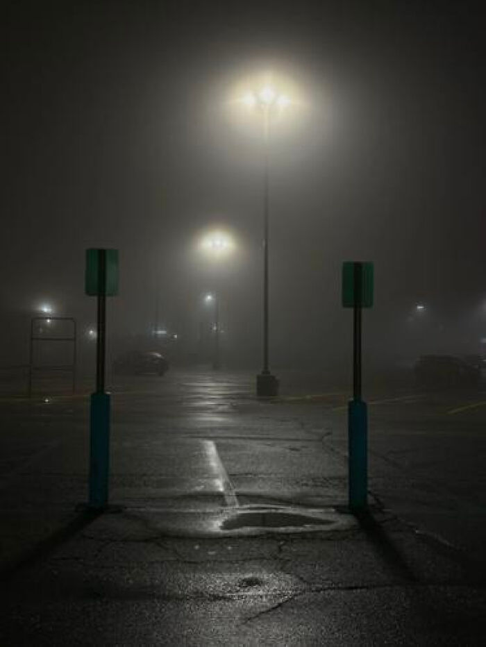 A foggy, dark parking lot illuminated by tall streetlights, evoking an unsettling liminal space.
