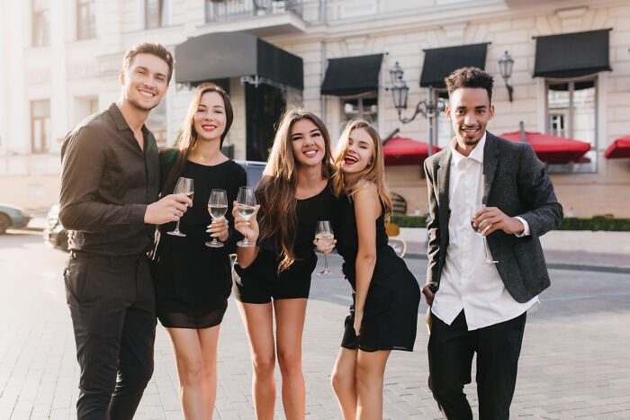 A group of friends in black attire, holding wine glasses and smiling on a city street, reflecting disturbing things people learned about their friends.