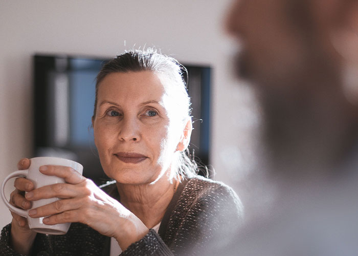 A woman with a serious expression holding a white mug, listening to someone off-camera, discussing a drunken tantrum.