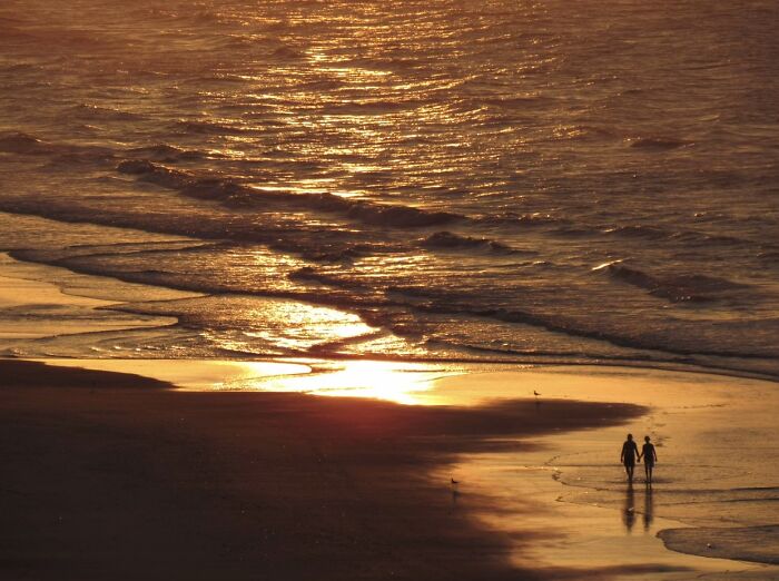 Two strangers walk hand-in-hand on a beach at sunset, embodying the wholesome "Like A Painting" photo trend.