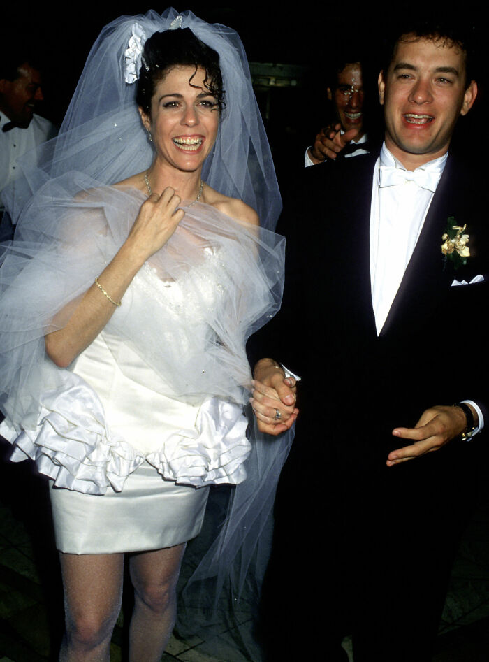 Rita Wilson in a short white wedding dress with a ruffled skirt and Tom Hanks in a tuxedo at their wedding.