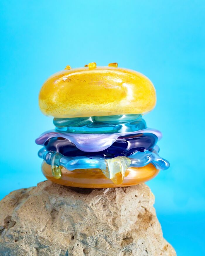 A hyper-realistic glass food burger with blue and purple layers, sitting on a rock against a blue background.
