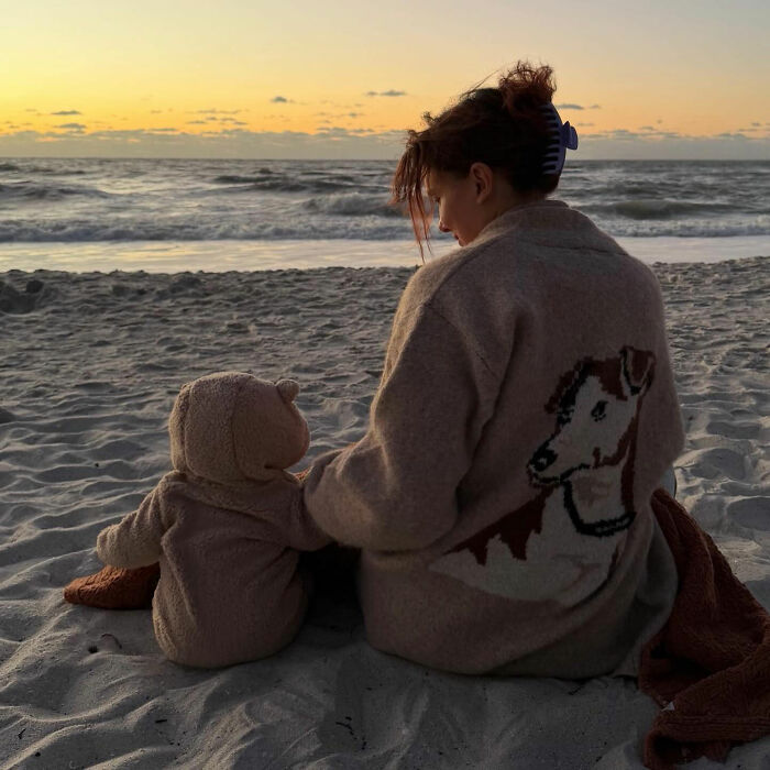 Millie Bobby Brown sitting on the beach next to a baby in a bear onesie at sunset