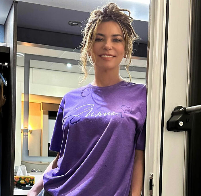 Shania Twain smiling in a purple t-shirt, showcasing her transformation and new beauty standards. Shania Twain smiling in a purple t-shirt, showcasing her transformation and new beauty standards.