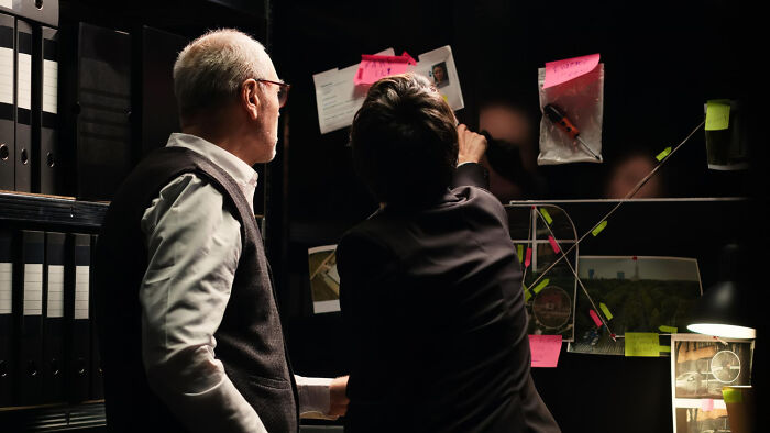Two detectives examining a wall board with photos and notes, working to solve unsolved mysteries.