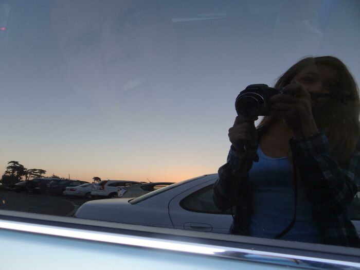 Reflection of a person holding a camera in a car window with sunset