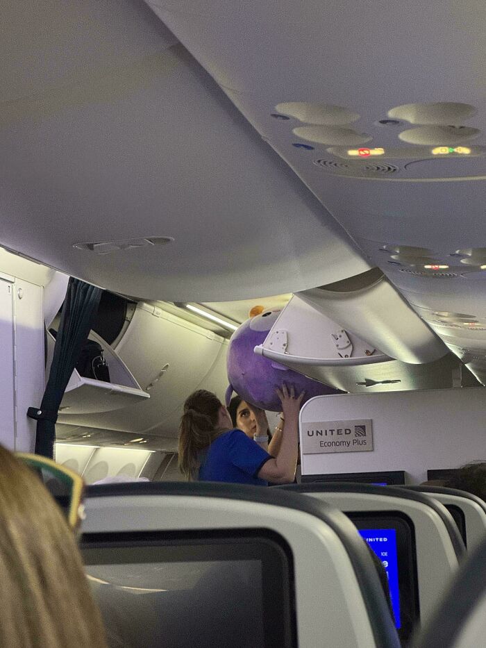A woman on an airplane struggling to fit an oversized purple stuffed animal into the overhead bin, showing airport chaos.
