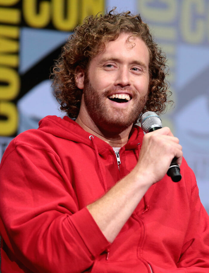 T.J. Miller speaking and smiling during a public event, wearing a red hoodie, showcasing his career highlights