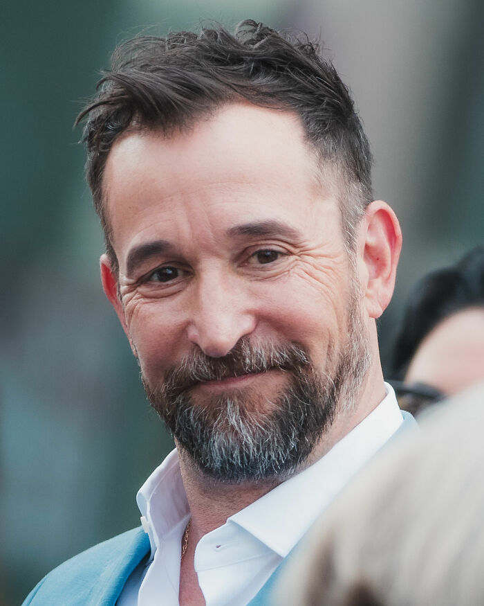 Close-up portrait of Noah Wyle smiling with beard and wearing a light blue suit jacket