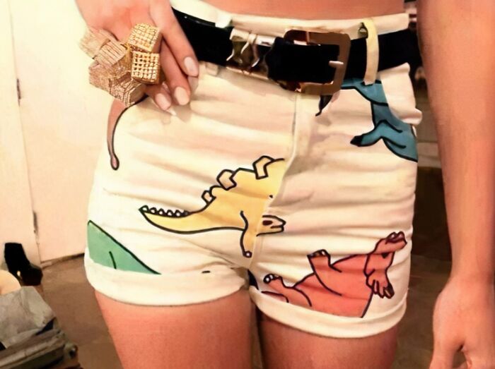 A person wearing white shorts with colorful dinosaur illustrations and a large gold ring, a questionable clothing design.