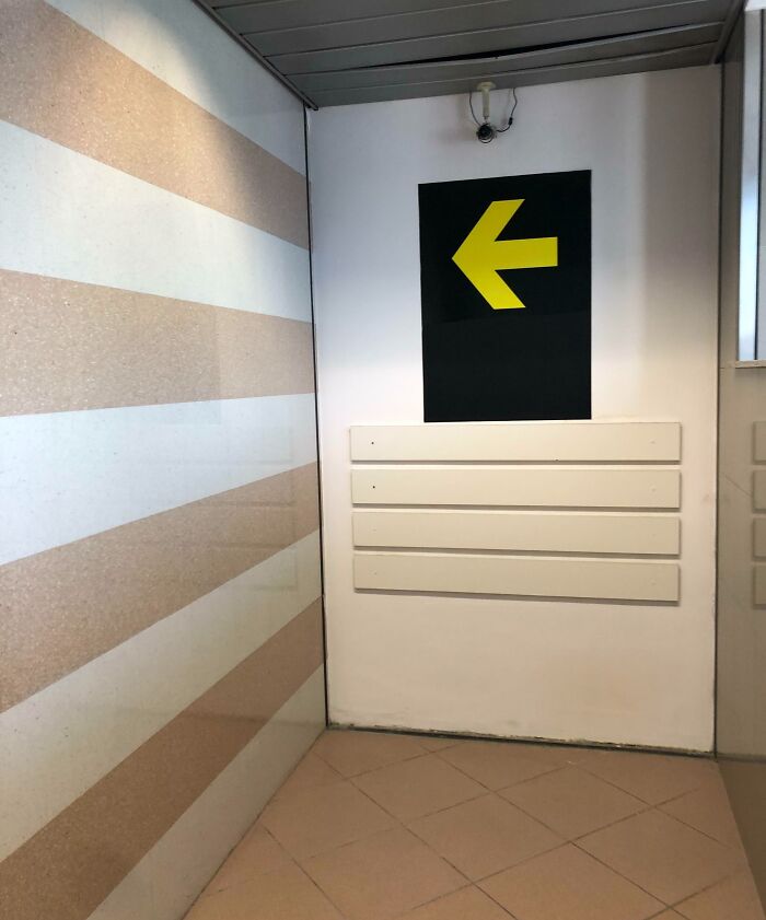 A yellow arrow on a black sign pointing left at an airport, highlighting chaotic airport navigation.