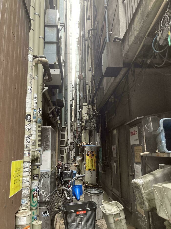 Narrow urban alley cluttered with bicycles, pipes, and wires between tall buildings