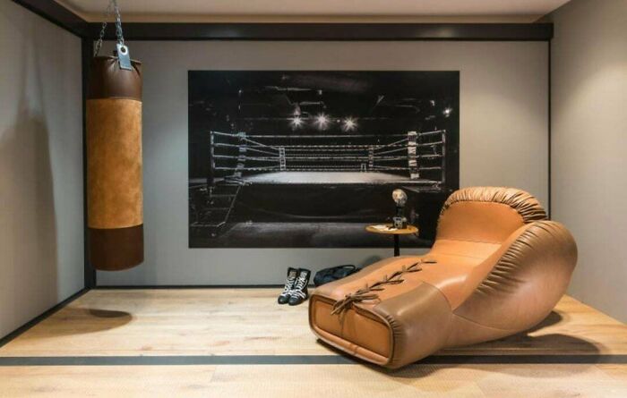 A room featuring a large brown boxing glove shaped couch next to a punching bag and a boxing ring picture, a bold interior designer statement.