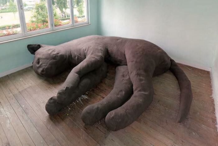 A giant, soft, dark gray cat-shaped couch lying on a wooden floor, an imaginative interior design piece.