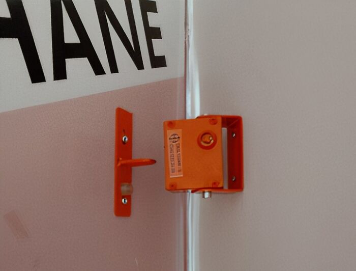 An orange metal object, possibly a security sensor or alarm component, mounted on a white wall near a corner, hinting at toxic teachers.