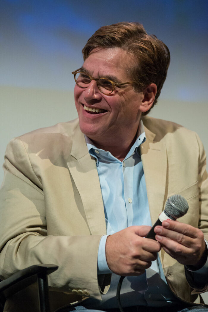 Aaron Sorkin speaking at an event holding a microphone and smiling, wearing glasses and a beige jacket