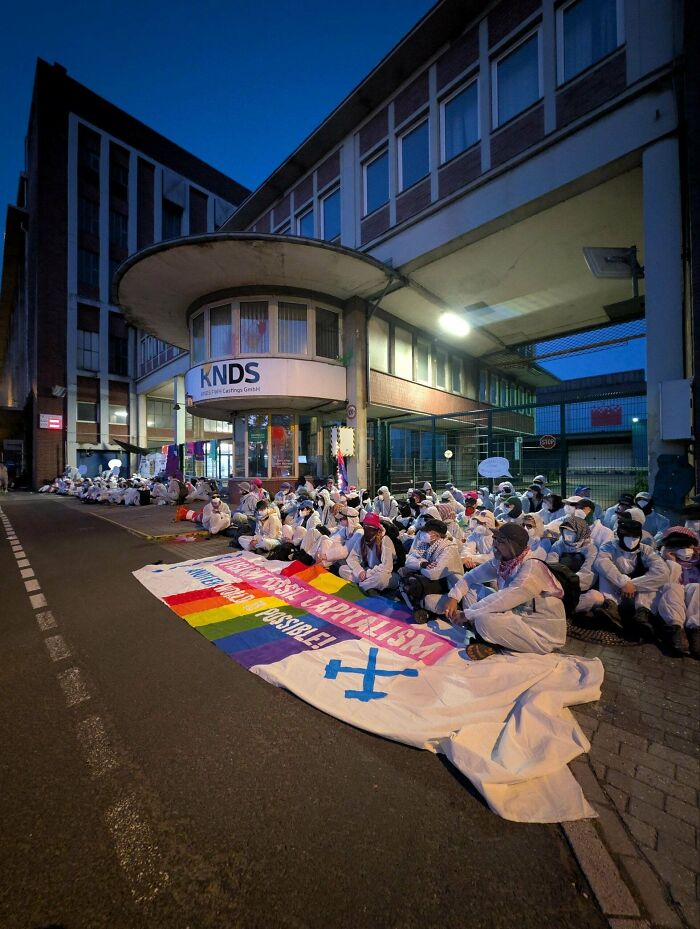 A group of protestors in white suits sitting outside a building with a large rainbow banner, showcasing a real life chaotic good action.