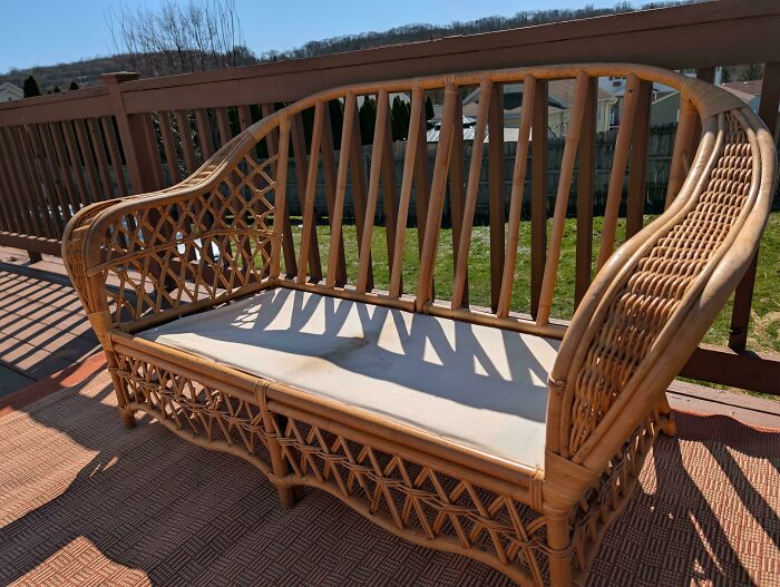 A light brown wicker bench with a light cushion sitting on a deck, a successful dumpster diving find.