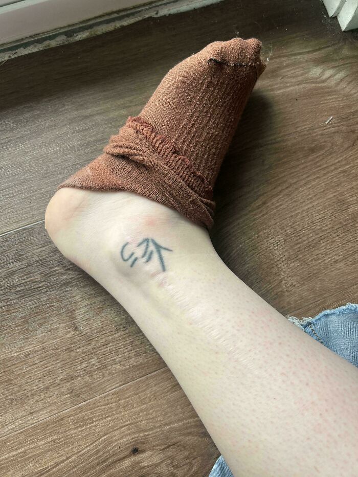 Mildly interesting pictures: An ankle with a small tattoo and a brown sock scrunched up on a wooden floor.